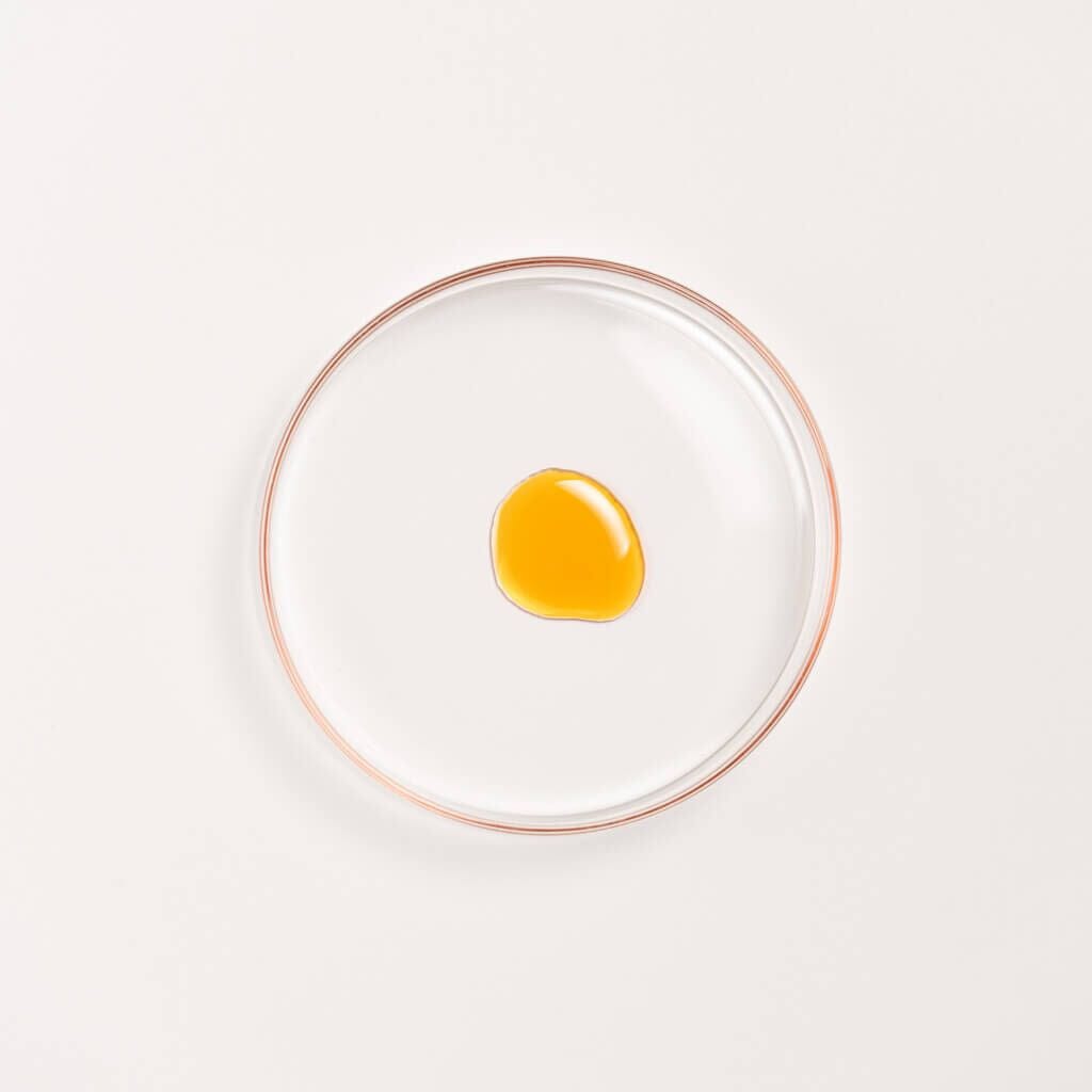 Manjish Glow Night Time Elixir texture: a drop of the amber oil in a round glass dish on a white background