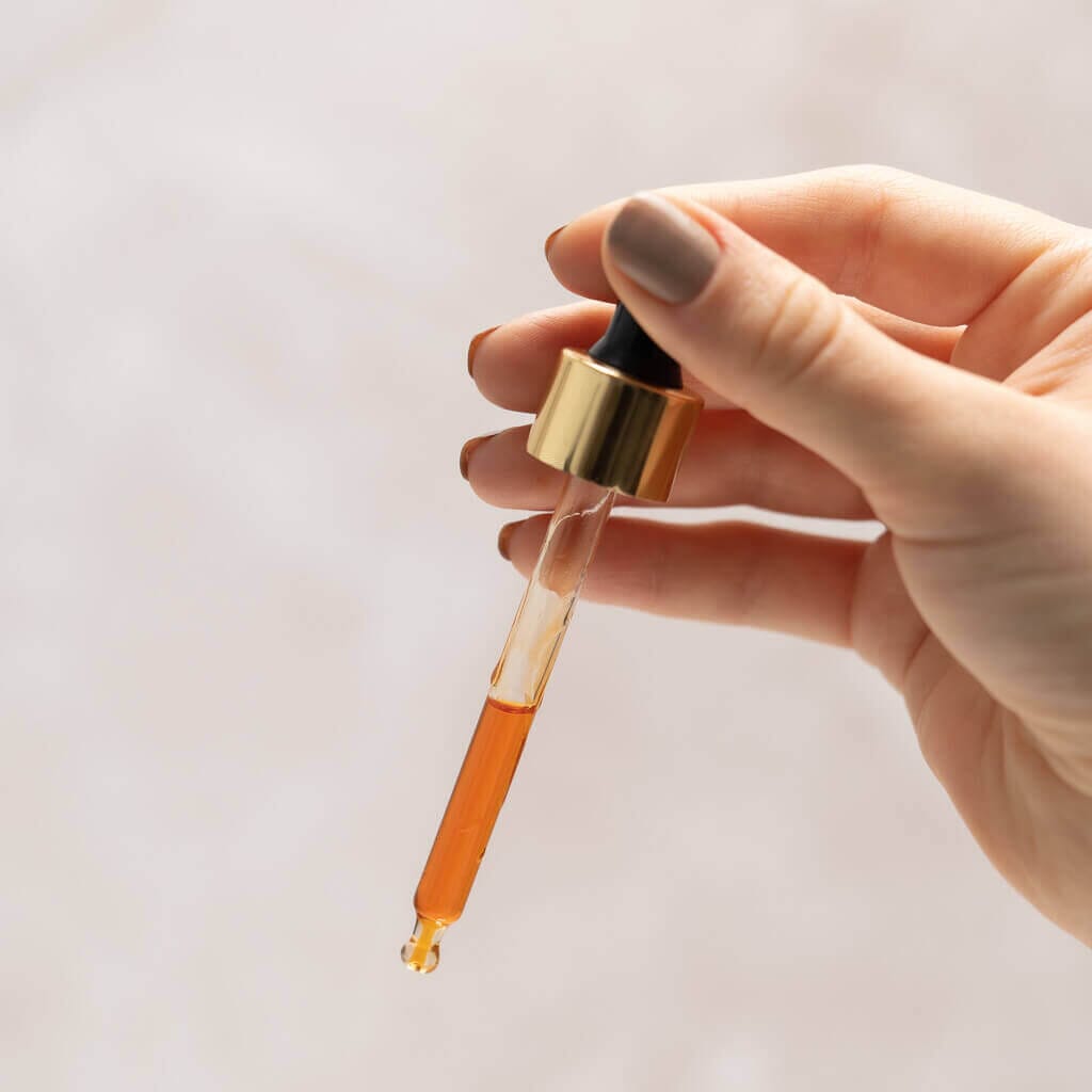 Manjish Glow Night Time Elixir: a hand holding the gold-collared dropper filled with orange oil, a drop at its tip, against a pale background