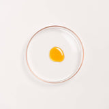 Manjish Glow Night Time Elixir texture: a drop of the amber oil in a round glass dish on a white background