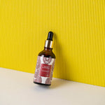 Manjish Glow Night Time Elixir bottle leaning against a yellow ribbed wall on a white surface