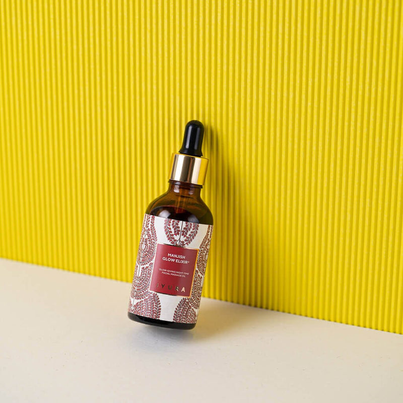Manjish Glow Night Time Elixir bottle leaning against a yellow ribbed wall on a white surface