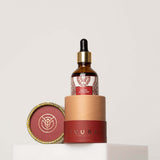 Manjish Glow Night Time Elixir: the dropper bottle standing in the base of its red tube on a white block, with the round lid, marked with the iYURA leaf logo, beside it