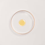 Kesaradi Daily Glow Oil texture: a drop of the pale golden oil in a glass dish with a rose-gold rim, on a white background