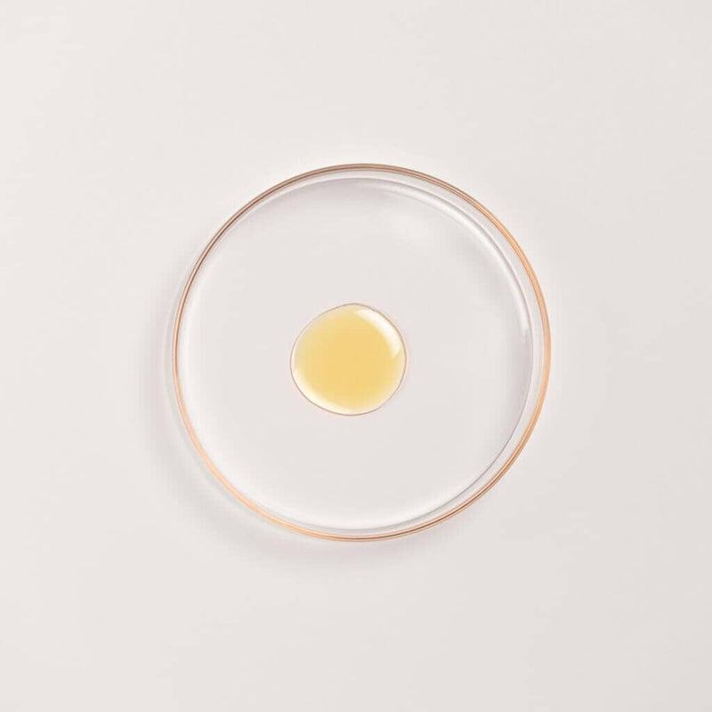 Kesaradi Daily Glow Oil texture: a drop of the pale golden oil in a glass dish with a rose-gold rim, on a white background